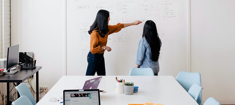 Two women pointing and directing their attention to a whiteboard with information in an office space.