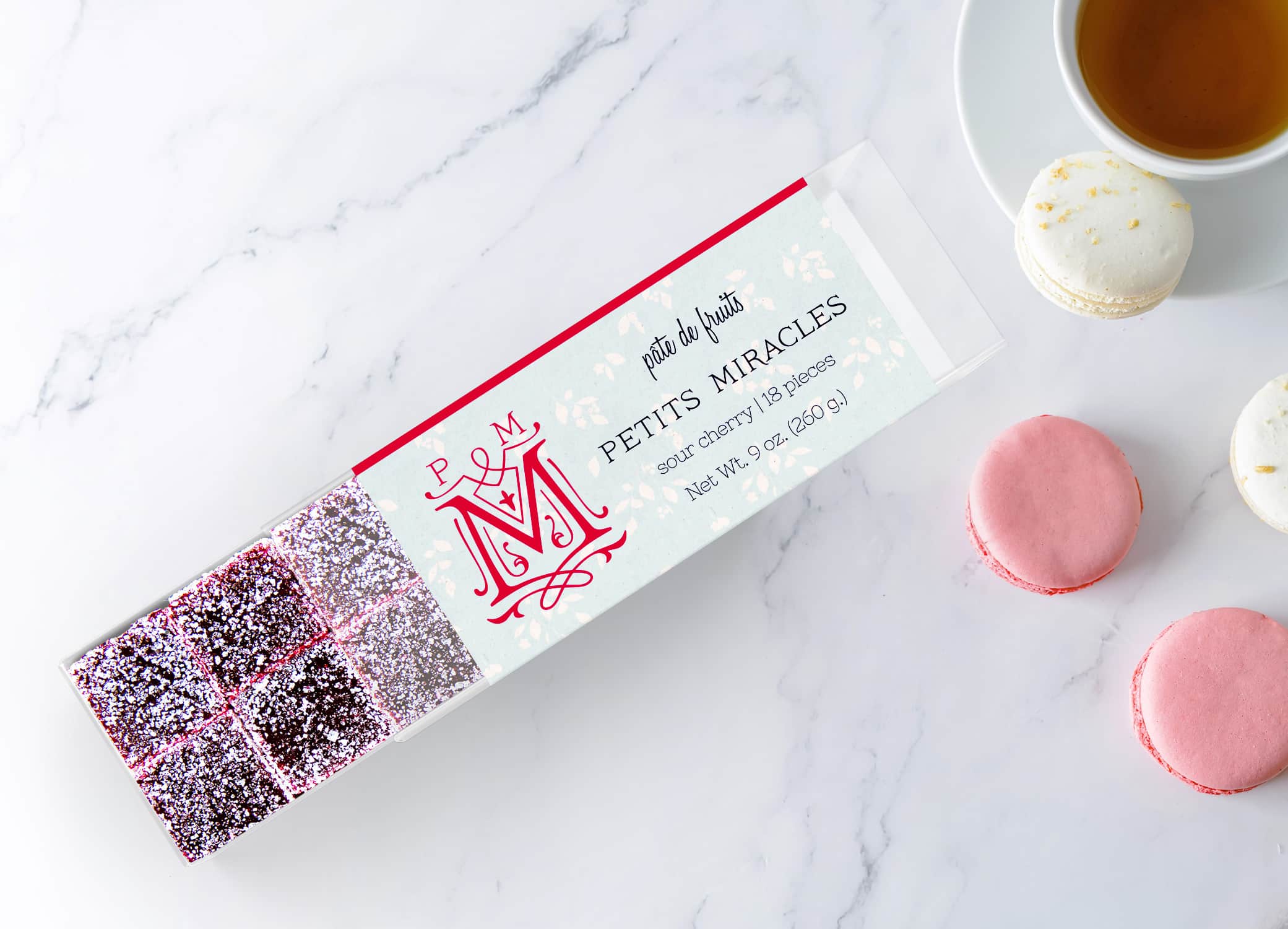 Petits Miracles Pate de Fruits packaging design with bright pink bands along the edges of the box and swirls around "M" icon next to type against a marble background with tea and macarons.