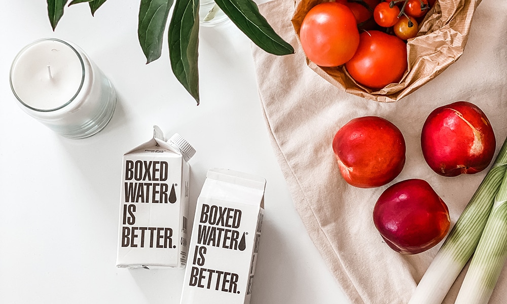Two boxes of water using paper packaging are placed among vegetables, fruits, plants, and a candle.